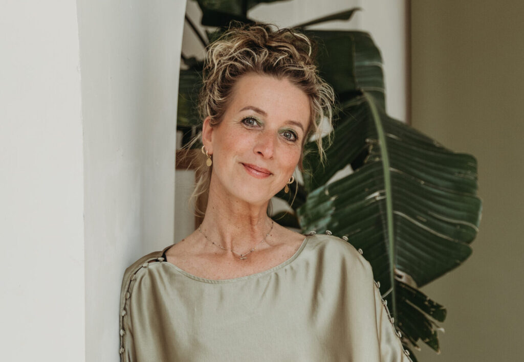 Woman with curly updo hairstyle wearing sage green satin blouse, leaning against white archway near tropical plant leaves